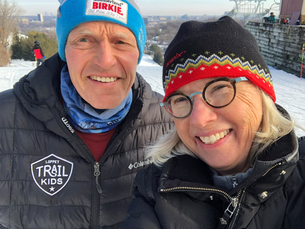 A close up selfie of John (left) and Gwen (right) bundled up in winter coats and hats with snow on the ground all around them.