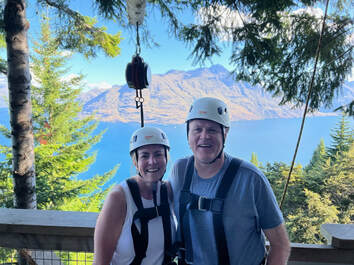 Meleah (left) and Chuck (right) stand on a walkway high in the trees, wearing ziplining safety gear (white helmets and harnesses), smiling in against a backdrop of a big blue lake and mountains.