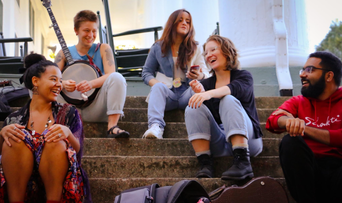 5 young adults of diverse backgrounds sit on stone steps, smiling and laughing with each other. Helen, in the back left, holds her banjo in her lap.