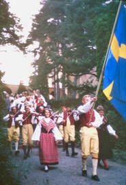 PictureMargit marches in a parade in Sweden in 1966. In front is a man waving a large Swedish flag; behind a small band marches playing violins. All are dressed in traditional clothing.