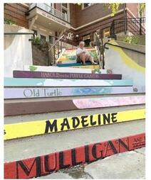 Kathy, an older white woman with white hair wearing a t-shirt and shorts, sits on the colorful steps to the Northfield Public Library. Each step is painted with a different book title and background, painted to resemble the book’s cover art.  From closest to furthest, some of the books are: “Madeline”, “Old Turtle”, and “Harold and the Purple Crayon”.