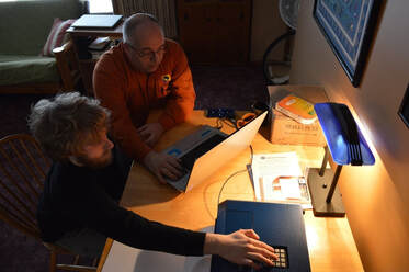 John Thomas sits in front of a laptop at a living room table, while Andrew Rossow sits next to him. Andrew has his hand on the embosser, while John helps him set it up.