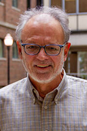 A close headshot of Peter Webb smiling; he is an older white man wearing a grey gingham button down shirt & blue-framed glasses.