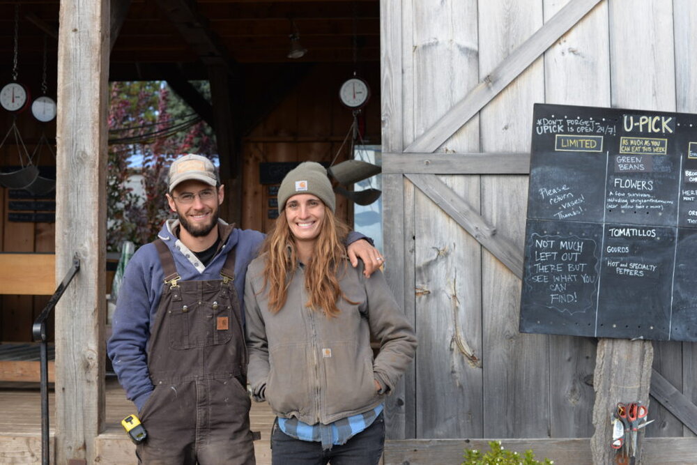 Andrew (left) and Betsy (right) stand outside the Spring Wind barn, dressed in light jackets and hats. The U-pick chalkboard peeks in from the right, which notes different crops that CSA members can pick themselves.